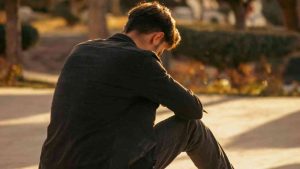 A man in dark clothing sits with his back to the camera, looking downward.