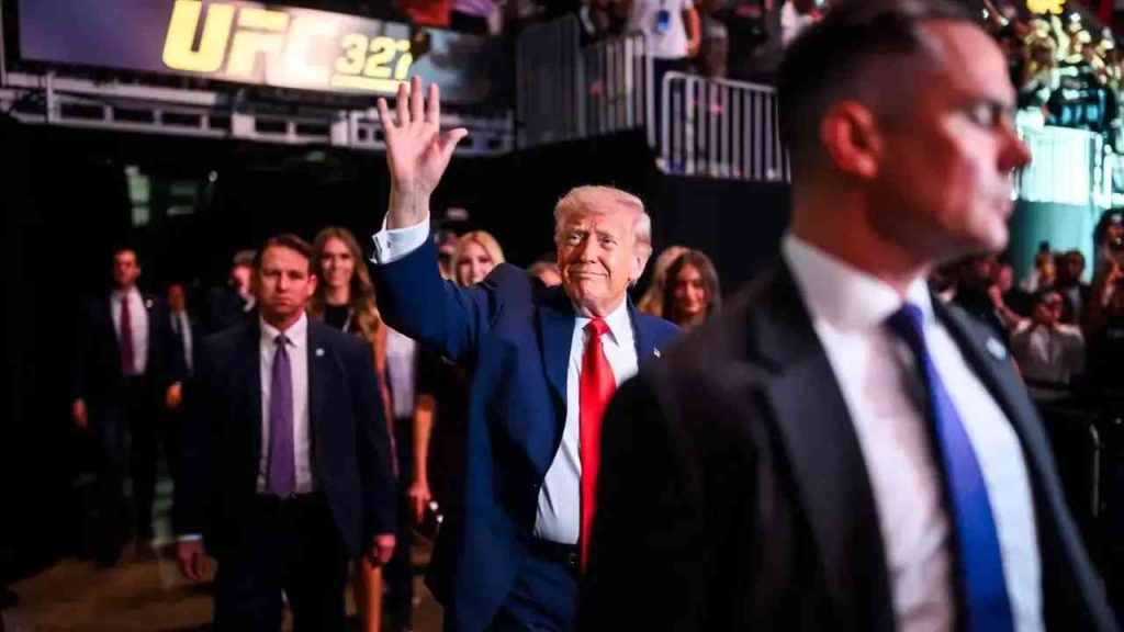 Donald Trump waves to the crowd during a UFC gala, surrounded by security and close associates. The image illustrates Trump’s political style, his relationship with voters, and the controversy surrounding his second term, the war with Iran, and the credibility of the “America First” slogan.