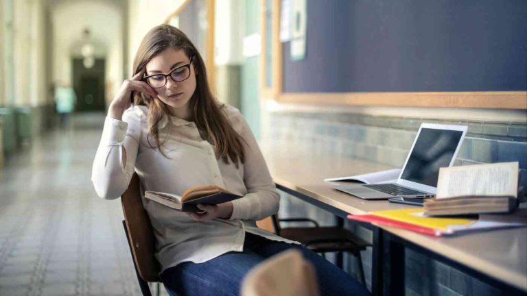 A schoolgirl is reading a book beside an open computer, wondering whether AI in schools is a good idea.