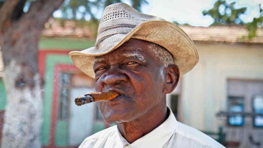 An older man in a straw hat smoking a cigar against a backdrop of buildings.