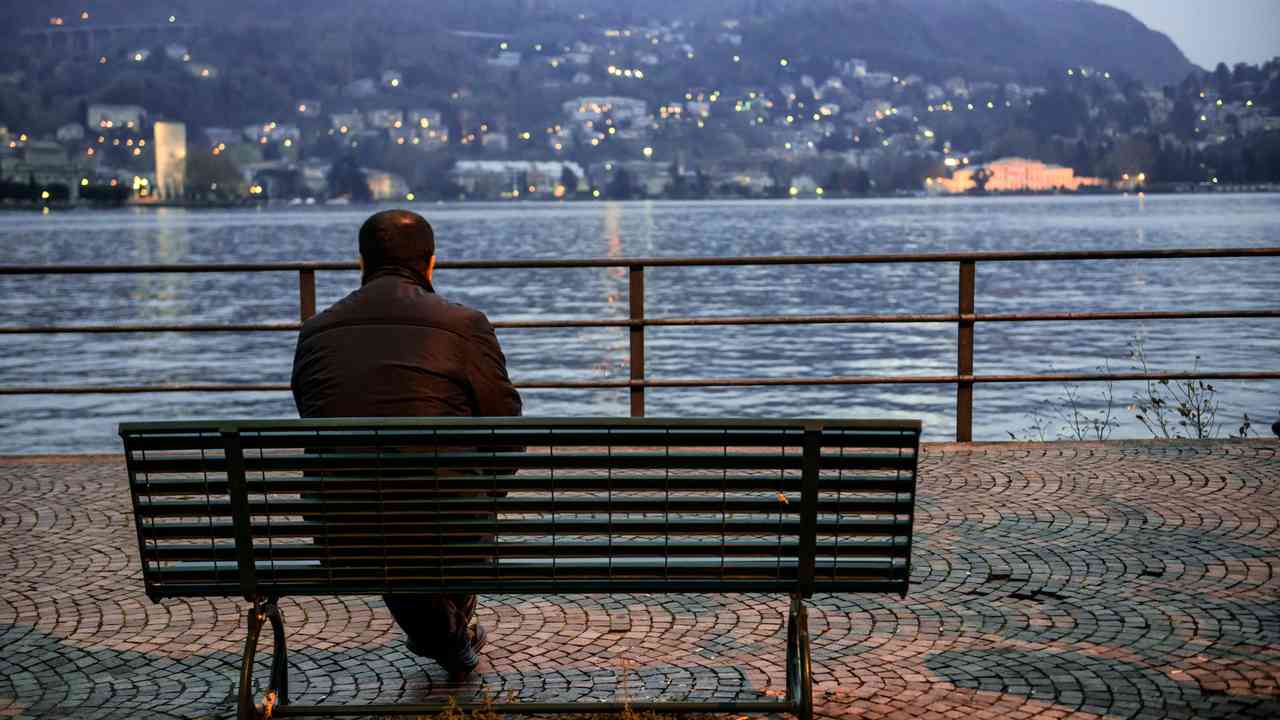 A lonely man sits on an empty bench by a lake, looking at the city lights.