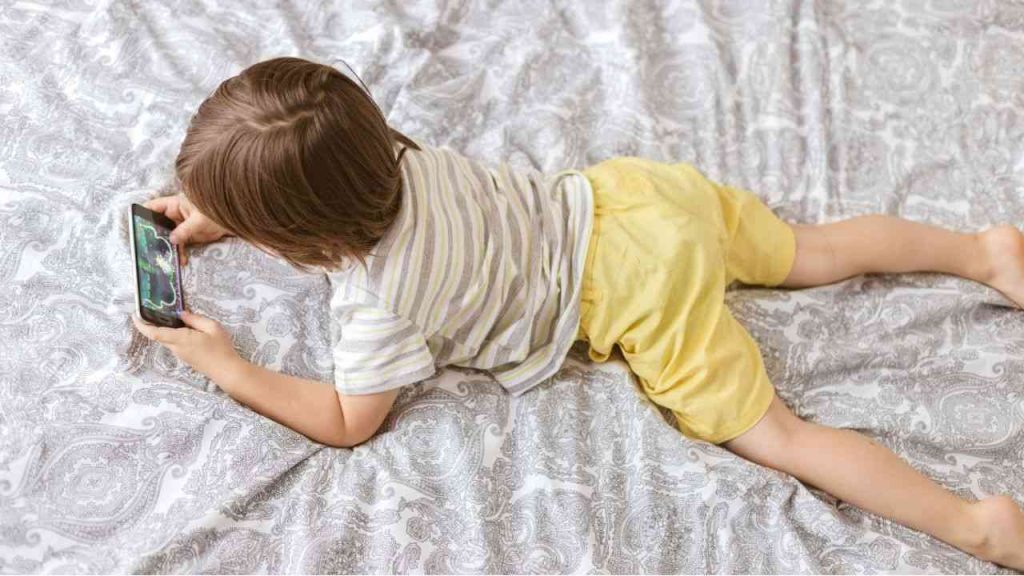 A child lies on a bedspread, playing a game on social media. The image illustrates the problem of children spending too much time on social media and children’s phone addiction.