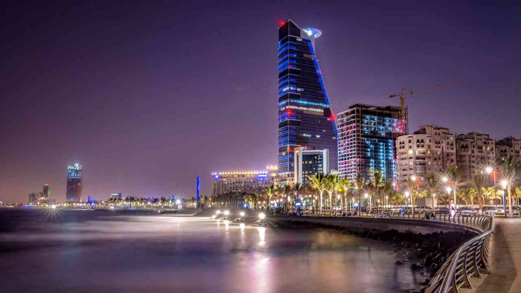 A nighttime view of Jeddah, Saudi Arabia, with modern skyscrapers and a waterfront promenade along the Red Sea.