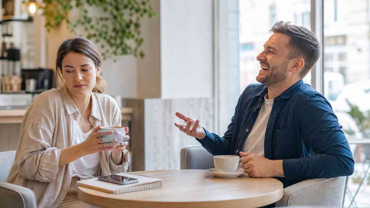 A smiling man jokes in a café, while the woman he is speaking with reacts with distance, showing different types of humor.
