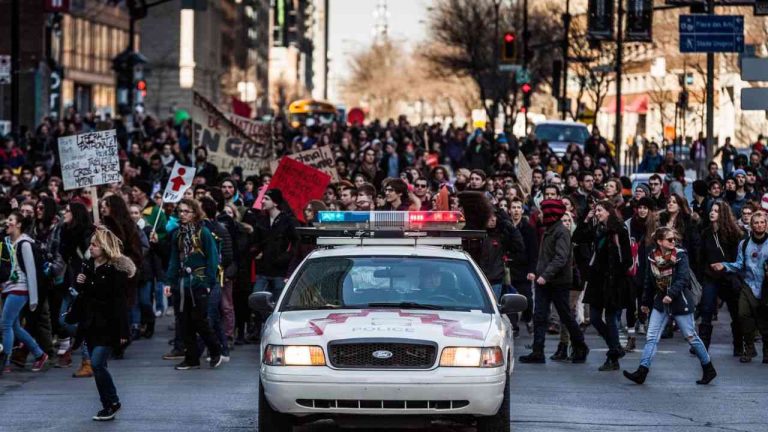 A police car drives along a road, followed closely by a crowd of people, illustrating the causes of political conflict.