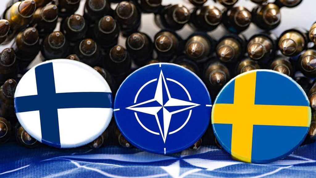 Finlandia zamyka granicę: trzy przypinki, Finlandii, NATO i Szwecji, w tle za nimi amunicja