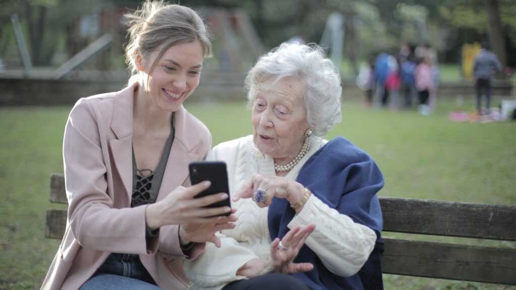 Starsi rodzice opieka. Na zdjęciu seniorka i młoda kobieta siedzą na ławce, młoda kobieta pokazuje seniorce coś w telefonie. Uśmiechają się