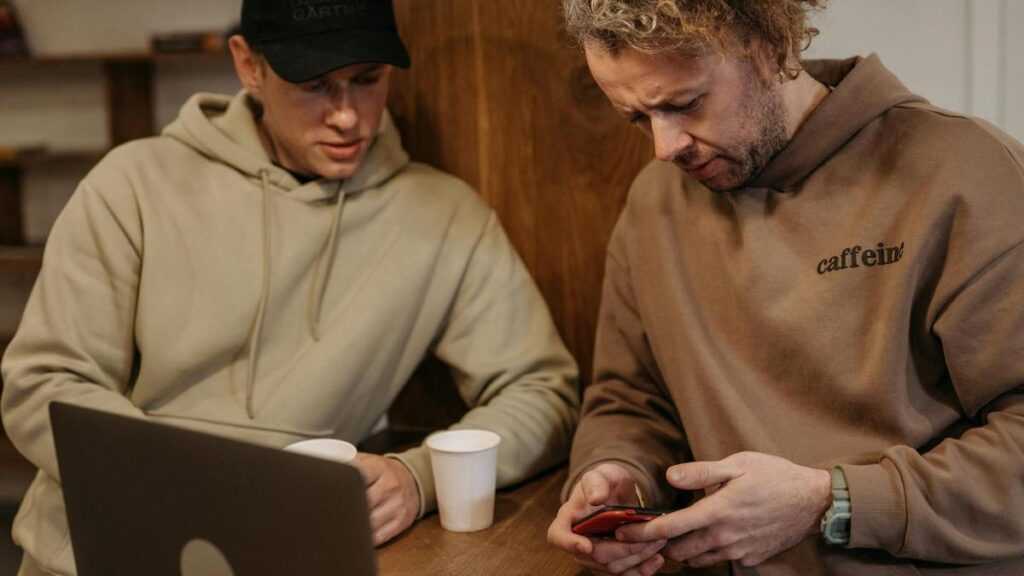 Iluzja wiedzy i sieć: dwóch mężczyzn w bluzach z kapturem przy stole z kawą i laptopem, jeden z mężczyzn dostrzega coś na ekranie smartfona
