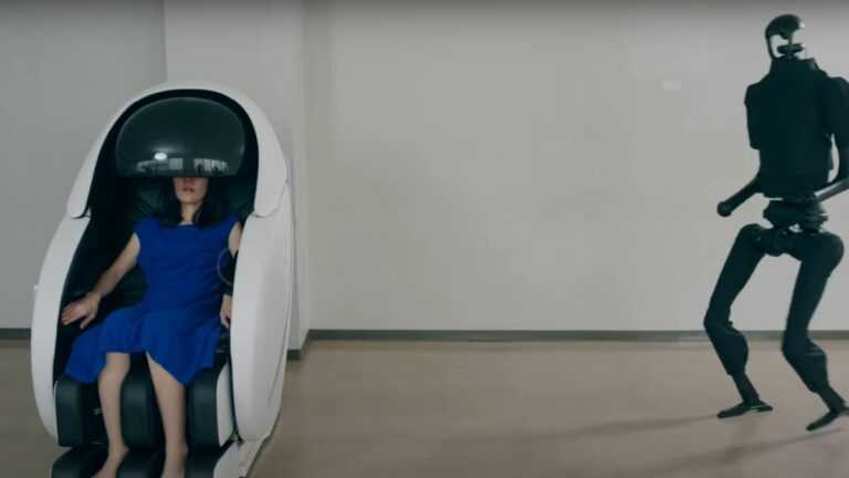 zdalnie sterowany robot, Fot. H2L Inc/Youtube, Kobieta siedzi w futurystycznej kapsule z zasłoniętą głową, podczas gdy humanoidalny robot zbliża się do niej w pustym pomieszczeniu.