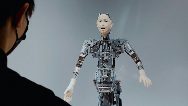 Zaufanie do AI: człowiek obrócony tyłem rozmawia z humanoidalnym robotem. Fot. Taiki Ishikawa / Unsplash
