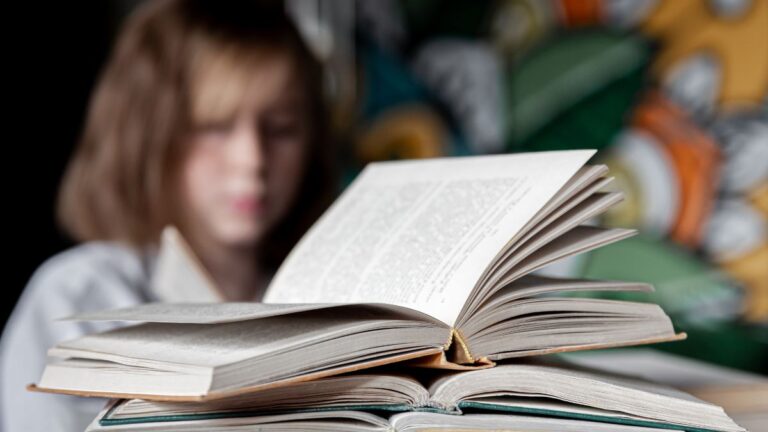 Dysleksja jest dziedziczna: na biurku stoi stos otwartych książek, w tle niewyraźna dziewczynka ze zmartwioną miną. Fot. pvproductions / freepik.