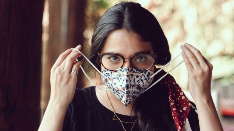 Koronawirus skutki uboczne, wpływ koronawirusa na mózg: kobieta w okularach zakłada maseczkę higieniczną. Fot. Andrés Salas / Unsplash