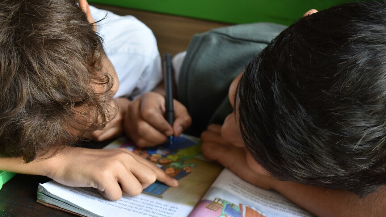 Dwoje uczniów odrabia obowiązkowe zadania domowe przy wspólnej książce, symbolizując zmiany w systemie edukacji.