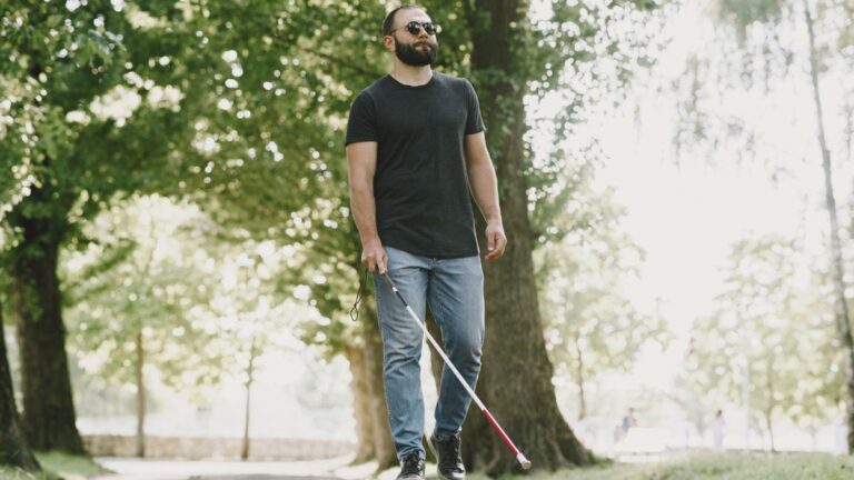 Przywrócenie wzroku niewidomym: niewidomy mężczyzna z okularami i laską idzie przez park. Fot. prostooleh / Freepik.