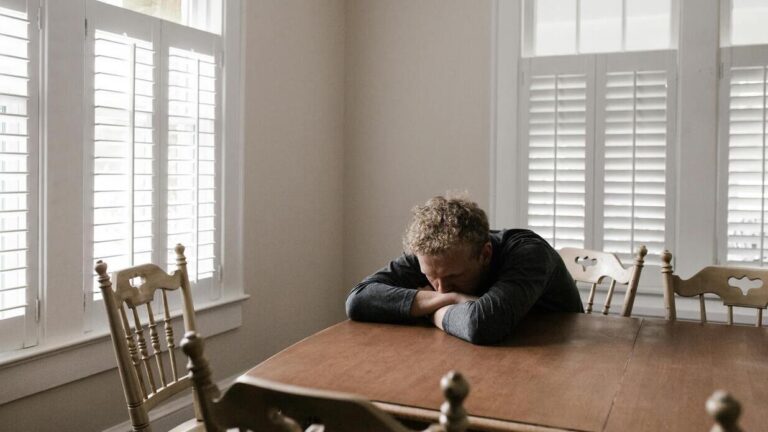 Mężczyzna siedzący samotnie przy stole z głową opartą na rękach, symbolizujący stres i niskie poczucie własnej wartości.