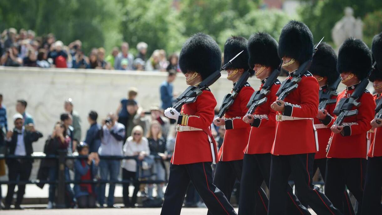 Zmiana warty przed pałacem w Londynie, żołnierze w czerwonych mundurach maszerują przed tłumem turystów.