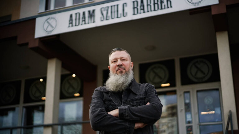 mężczyzna z brodą przed wejściem do barbershopu, postawa z założonymi rękami