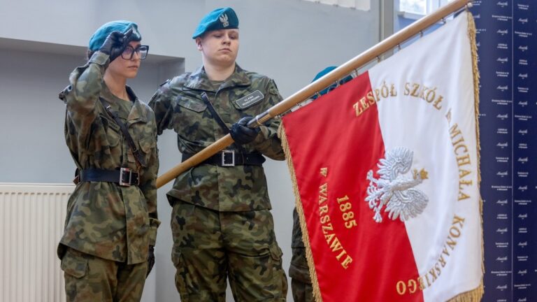 Uczniowie w mundurach podczas ceremonii w szkole mundurowej, salutujący przy sztandarze.