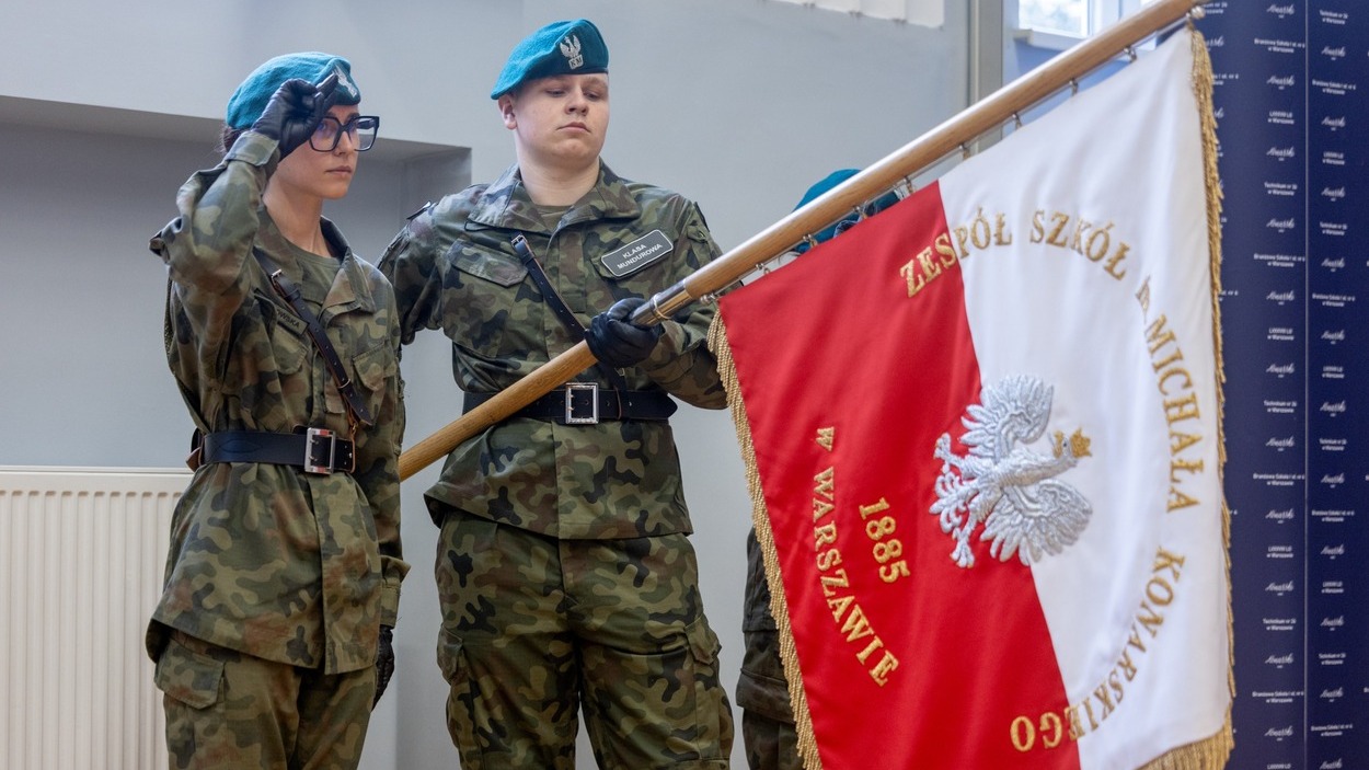 Uczniowie w mundurach podczas ceremonii w szkole mundurowej, salutujący przy sztandarze.