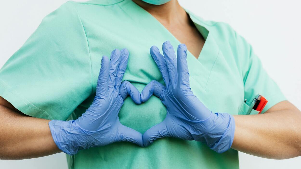 Lekarz w zielonym uniformie tworzy serce z dłoni w niebieskich rękawiczkach — symbol troski i nowoczesnej medycyny, w której małe roboty medyczne pomagają i leczą serce.