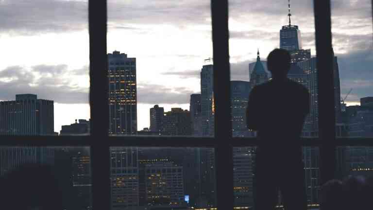 Zamyślony człowiek stojący przy oknie i patrzący na miasto o zmierzchu – symbol kryzysu współczesnego człowieka w świecie technologii