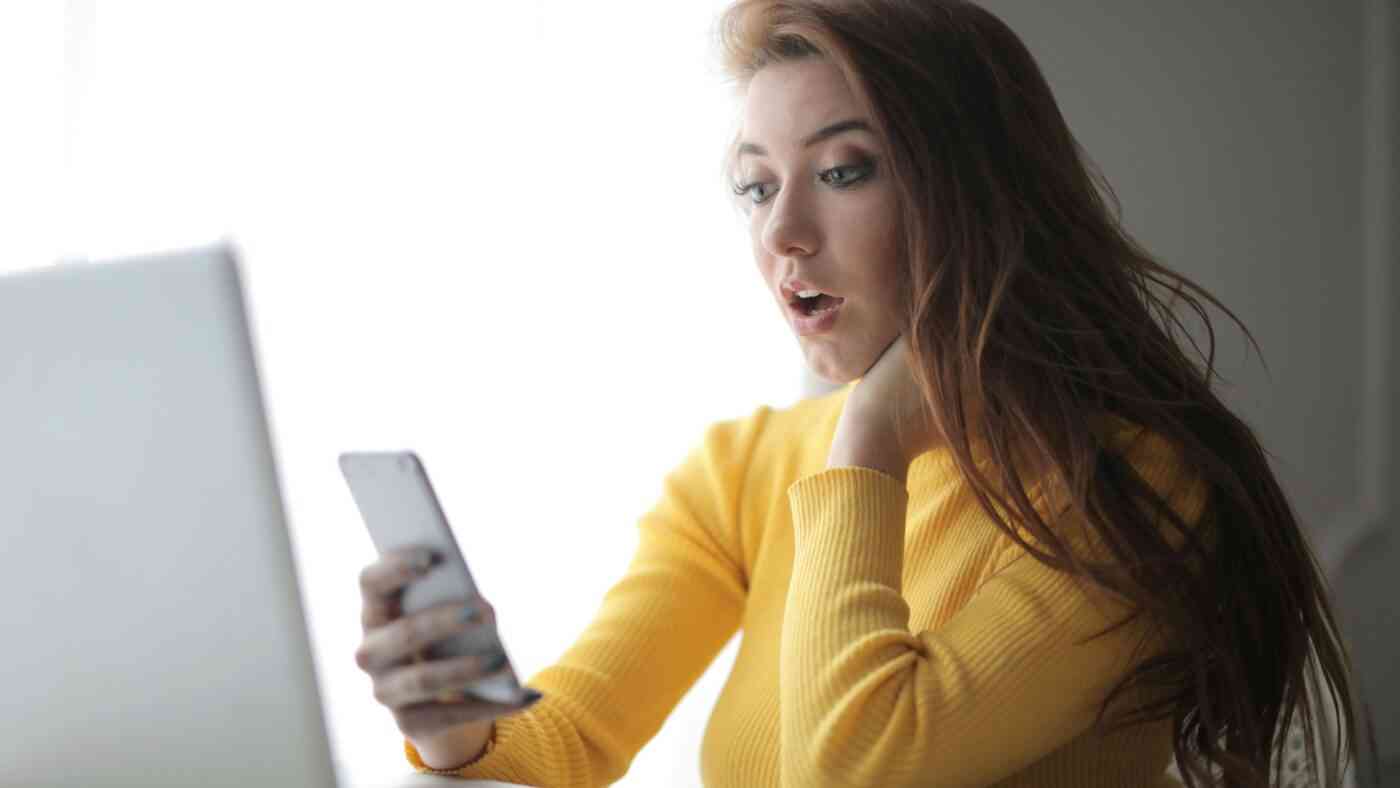 Kobieta w długich włosach i w żółtym swetrze patrzy na telefon i laptopa