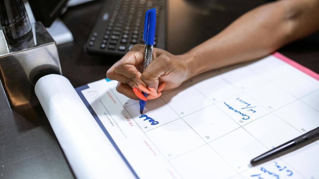 Osoba zaznaczająca datę na papierowym kalendarzu biurowym niebieskim markerem, planując wolne dla pracowników.