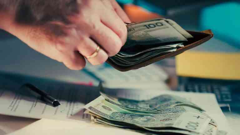 zbliżenie na rękę, która trzyma ciemny portfel i przelicza banknoty, które leżą na stole