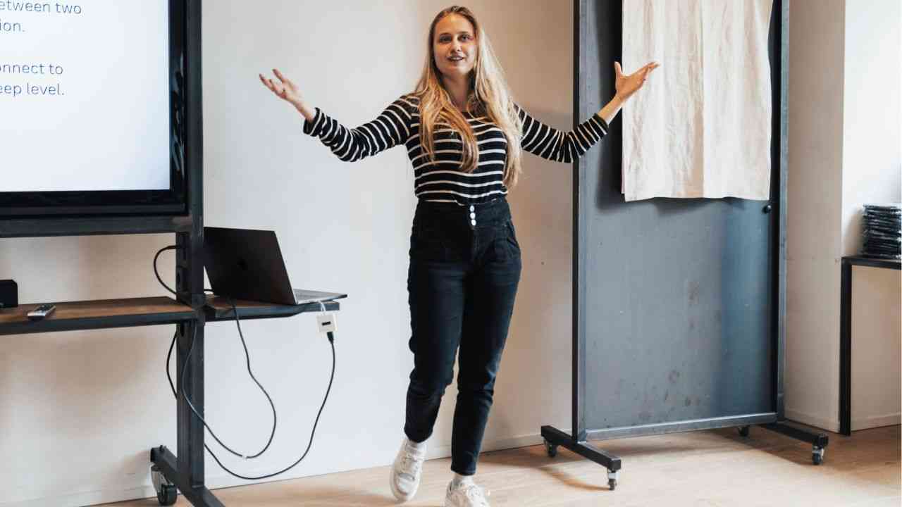 blondynka stoi przed tablicą z rozłożonymi rękami ubrana w czarne jeansy i bluzkę z czarno-białymi pasami