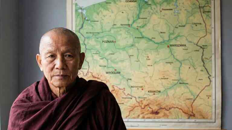 Starszy mnich buddyjski w bordowej szacie stoi na tle fizycznej mapy Polski, patrząc w obiektyw. Obraz ilustruje temat buddyzm w Polsce.