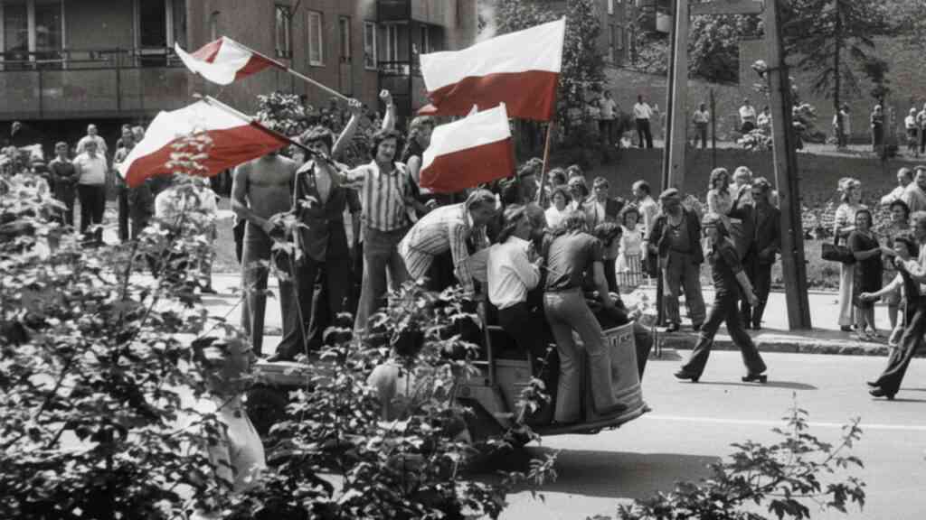 Grupa protestujących stoi na ciężarówce z biało-czerwonymi flagami podczas robotniczych demonstracji w PRL; wokół zgromadzony tłum mieszkańców obserwuje wydarzenia na ulicy w czerwcu 1976 roku w Radomiu.