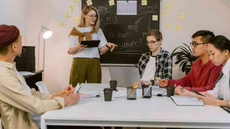 grupa studentów siedzi przy stole, kobieta stoi przed tablicą