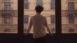 Młoda kobieta stojąca samotnie przy dużym oknie w deszczowy dzień, patrząca na miasto; nastrój podkreśla temat samotności i wpływu związku na psychikę.