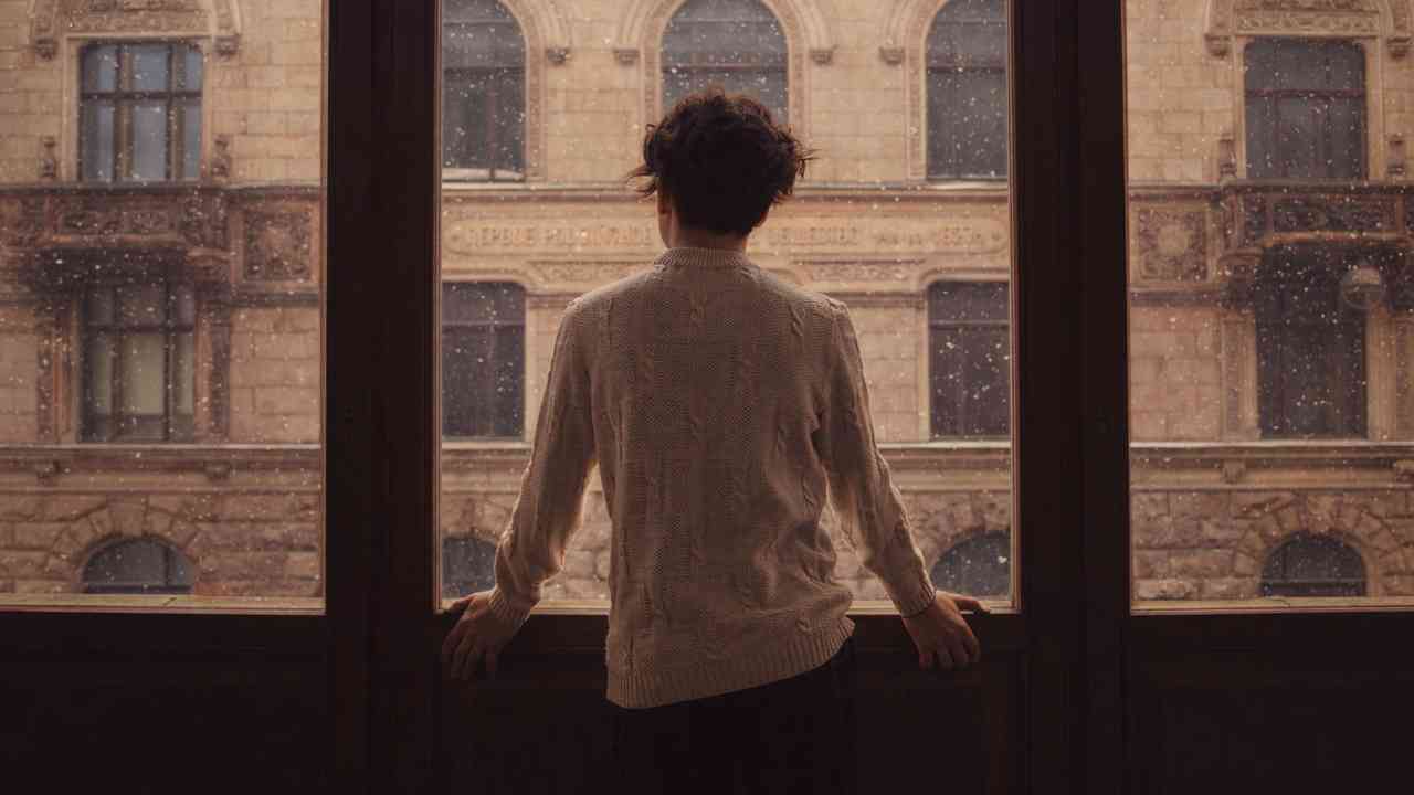 Młoda kobieta stojąca samotnie przy dużym oknie w deszczowy dzień, patrząca na miasto; nastrój podkreśla temat samotności i wpływu związku na psychikę.