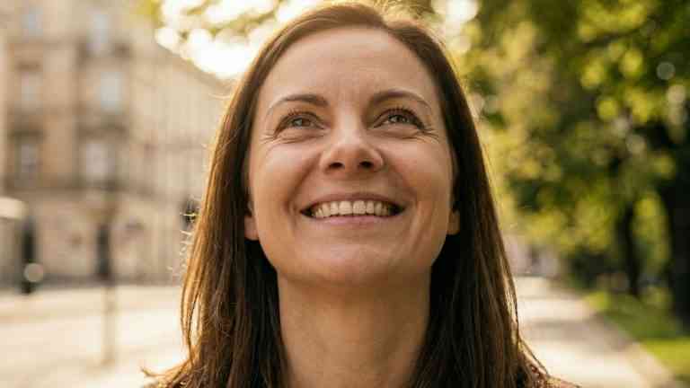 Portret uśmiechniętej kobiety spoglądającej z nadzieją w stronę słońca w miejskim parku, ilustrujący docenianie małych rzeczy.