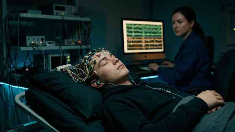 Młody mężczyzna podłączony do aparatury EEG w ciemnym pokoju pokazuje, jak kontrolować sny dzięki nowoczesnej technologii.