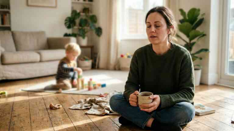 Kobieta w chwili wytchnienia z kubkiem napoju siedzi na podłodze obok bawiącego się w tle dziecka – wystarczająco dobra mama.