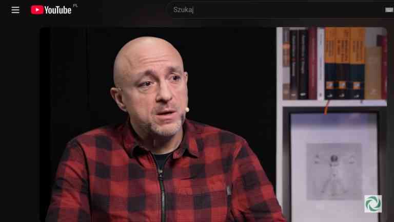 Przemysław Wójtowicz, snajper, zamyślony w czerwonej koszuli w kratę, siedzi podczas wywiadu na YT, patrząc w bok.