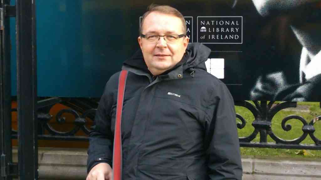 Piotr Gociek. Mężczyzna w czarnej kurtce pozuje na tle ciemnej ściany