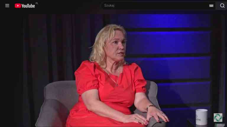 Prof. Klaudia Cymanow-Sosin w czerwonej sukience, siedząca w szarym fotelu podczas wywiadu na temat wpływu technologii na najmłodszych i relacji dzieci a ekrany. W tle studio z niebieskim oświetleniem.