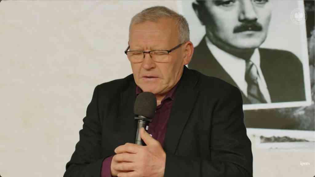 Bogdan Musiał w czarnym garniturze siedzi i trzyma mikrofon