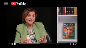 Aleksandra Ciejek, psycholog pozytywna, w zielonej marynarce podczas wywiadu wideo na platformie YouTube. W tle na białym regale z książkami stoi wyeksponowany magazyn z logo Holistic.
