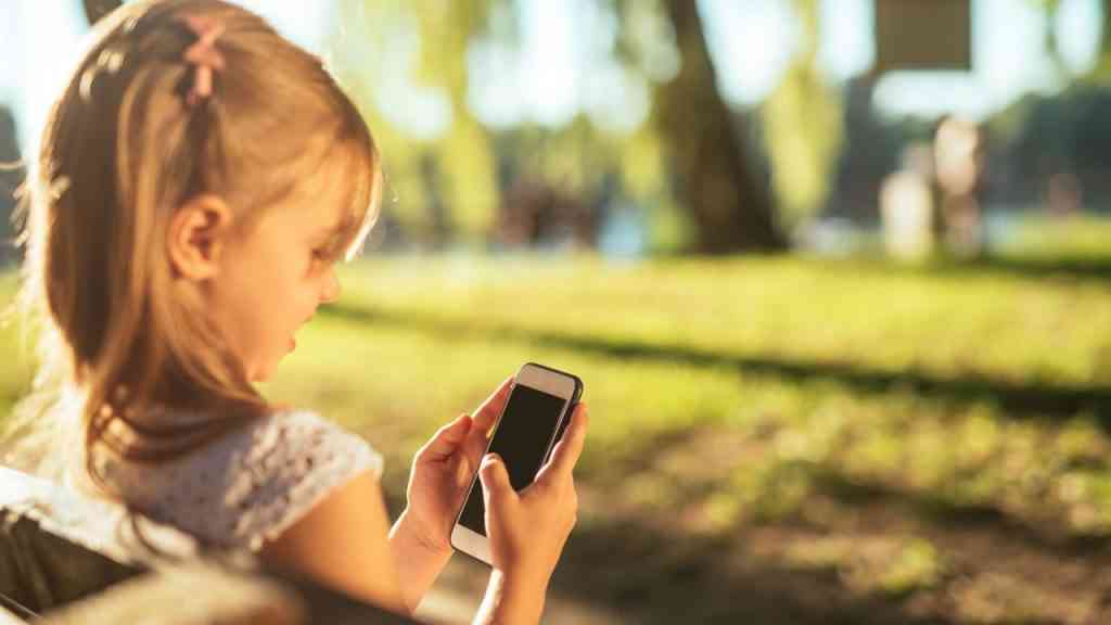 mała dziewczynka siedzi na ławce i trzyma telefon przed twarzą, co pokazuje zjawisko dzieci i mediów społecznościowych