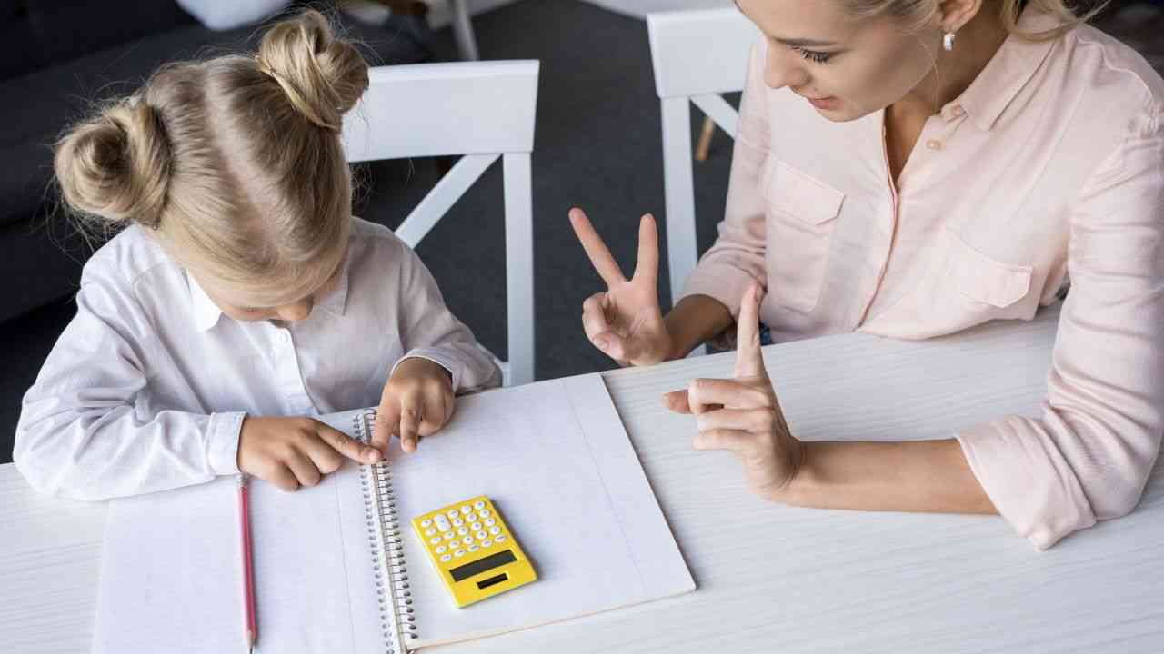 dwie osoby ubrane na biało siedzą przy białym stole, kobieta pomaga dziecku w problemach z matematyką
