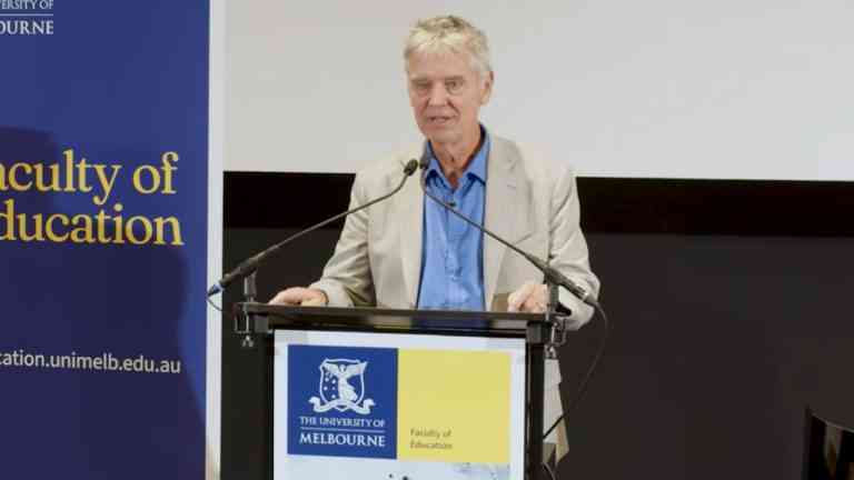 Profesot John Hattie ubrany w jasną marynarkę i niebieską koszulę mówi z mównicy, jak rozpoznać dobrą szkołę