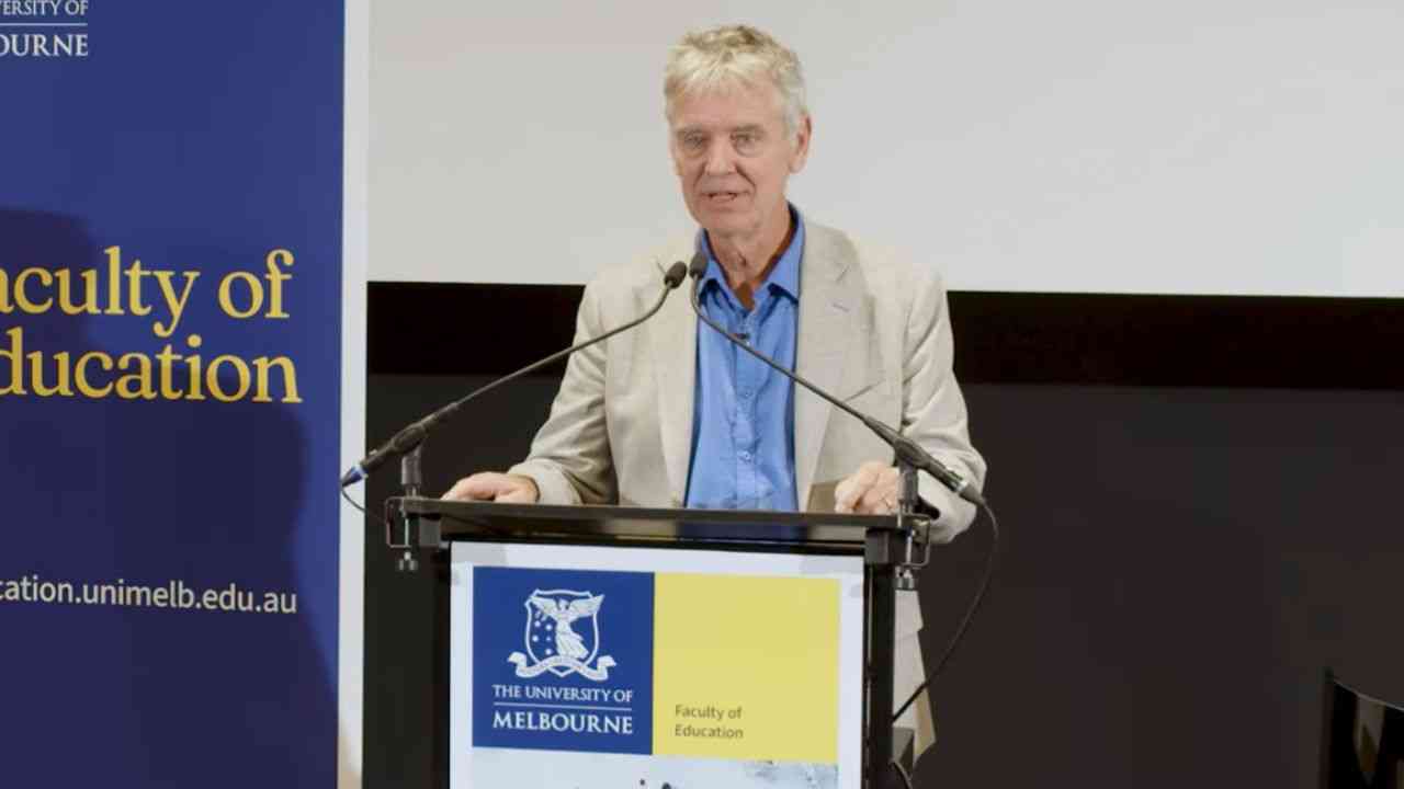 Profesot John Hattie ubrany w jasną marynarkę i niebieską koszulę mówi z mównicy, jak rozpoznać dobrą szkołę